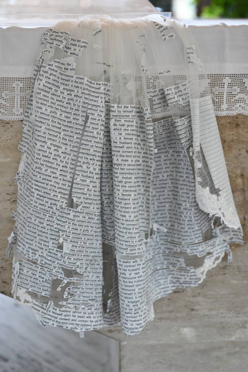 Wedding gown draped over stone, its skirt entirely papered in marital-rape legal text that frays like book pages