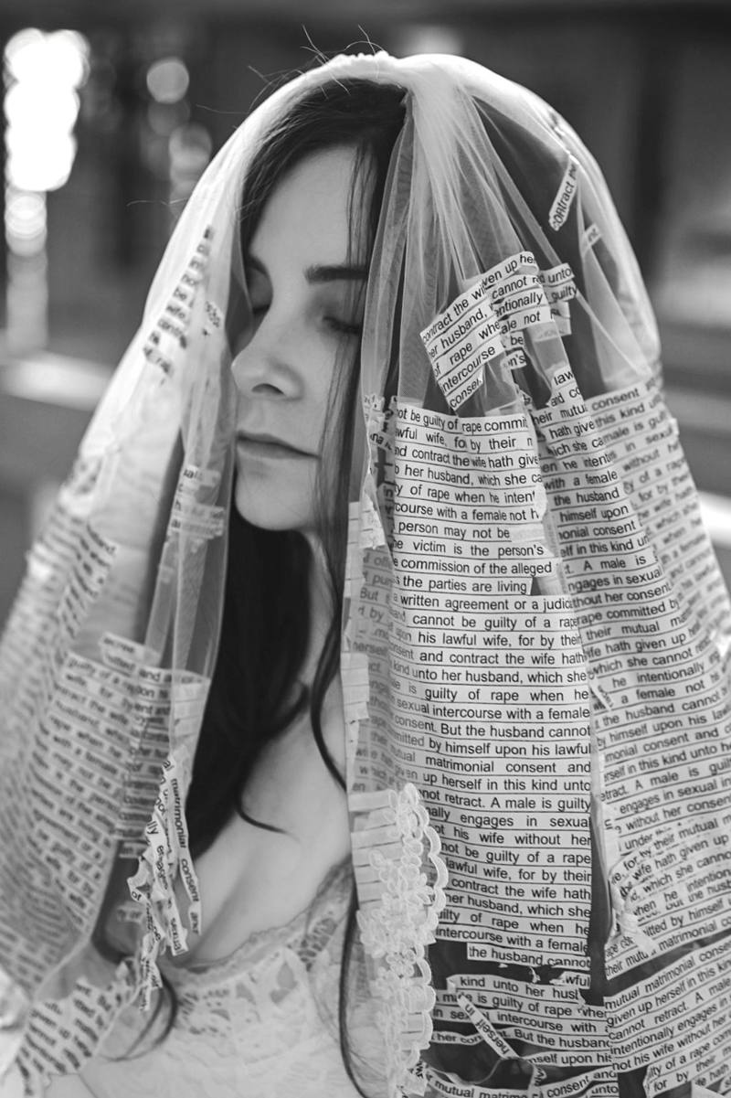 The veiled bride with eyes closed, strips of legal text peeling from the veil