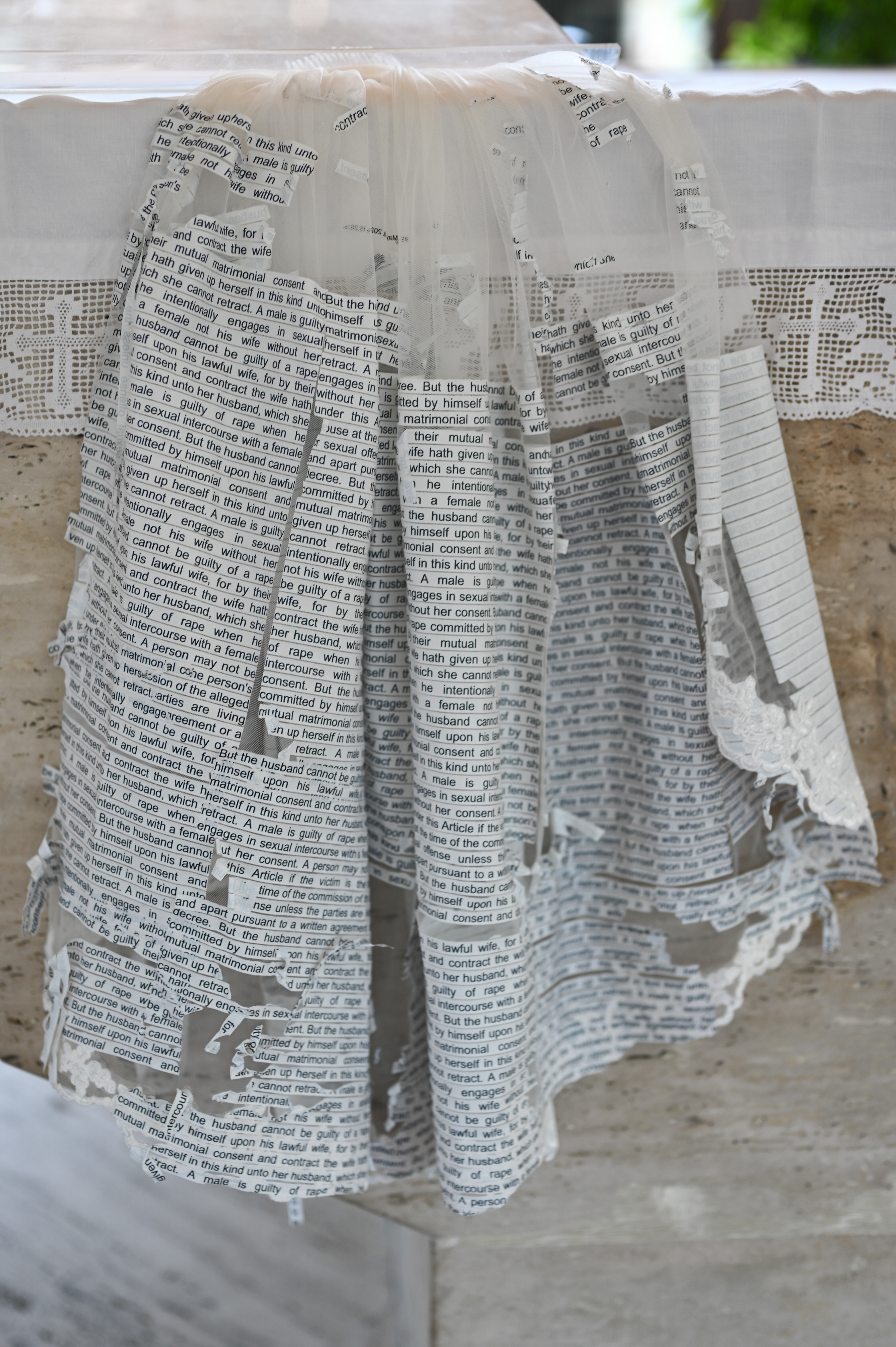 Wedding gown draped over stone, its skirt entirely papered in marital-rape legal text that frays like book pages